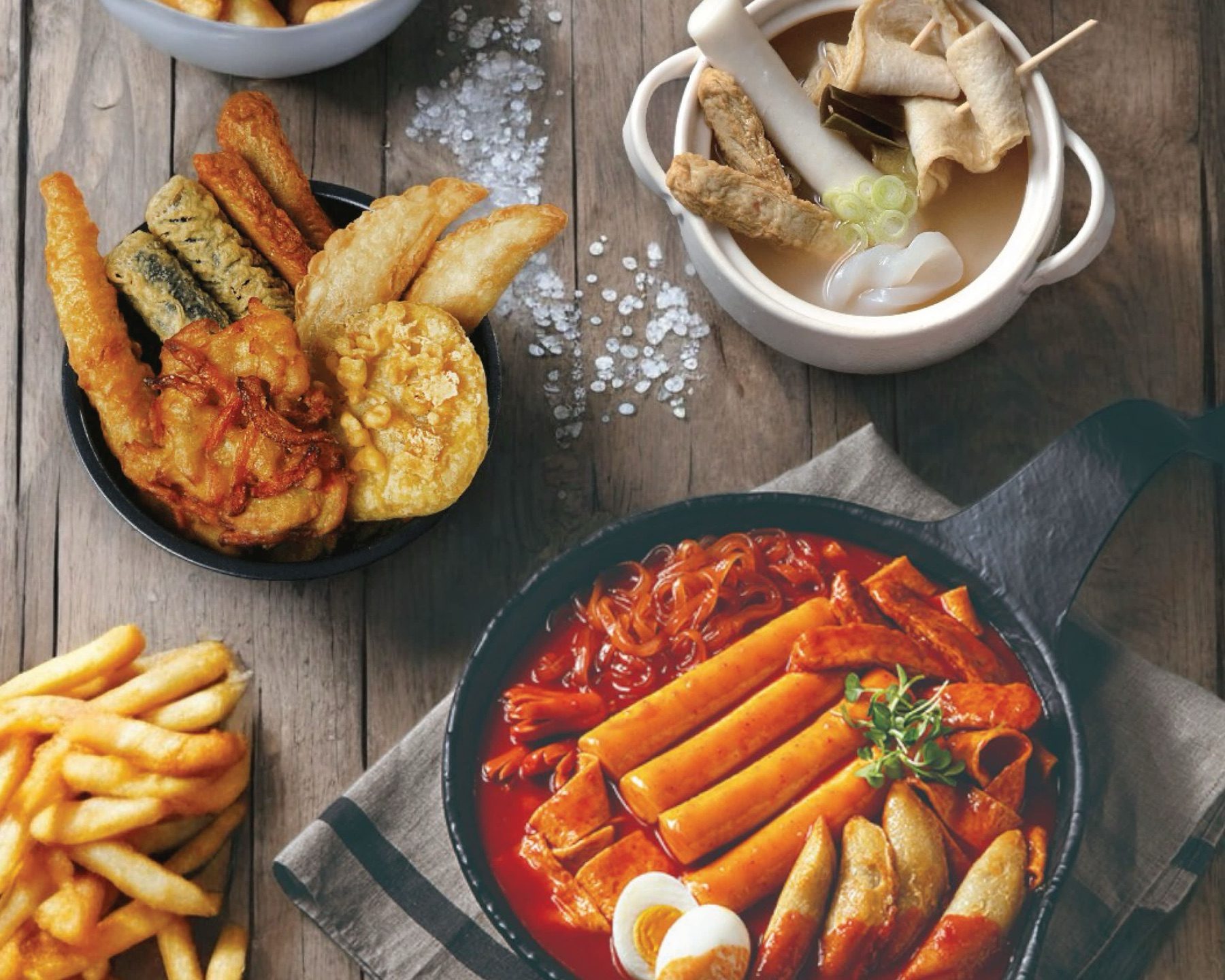A shared spread on a wooden table with a pan of jumbo tteokbokki, tempura and fish cake soup